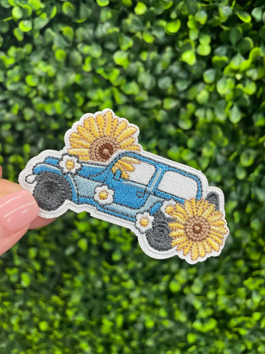 Embroidered patch of a blue car with sunflowers against a green leafy background