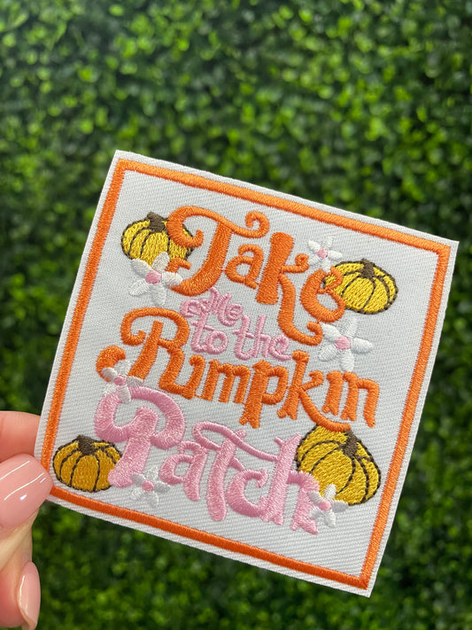 Embroidered patch featuring orange and pink text "Take me to the pumpkin patch" with pumpkin and flower designs around it, held against a green leafy background